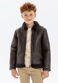 Veste bomber en faux cuir marron foncé avec un col en fourrure, fermeture zippée et deux poches avant. Portée sur un pull crème.