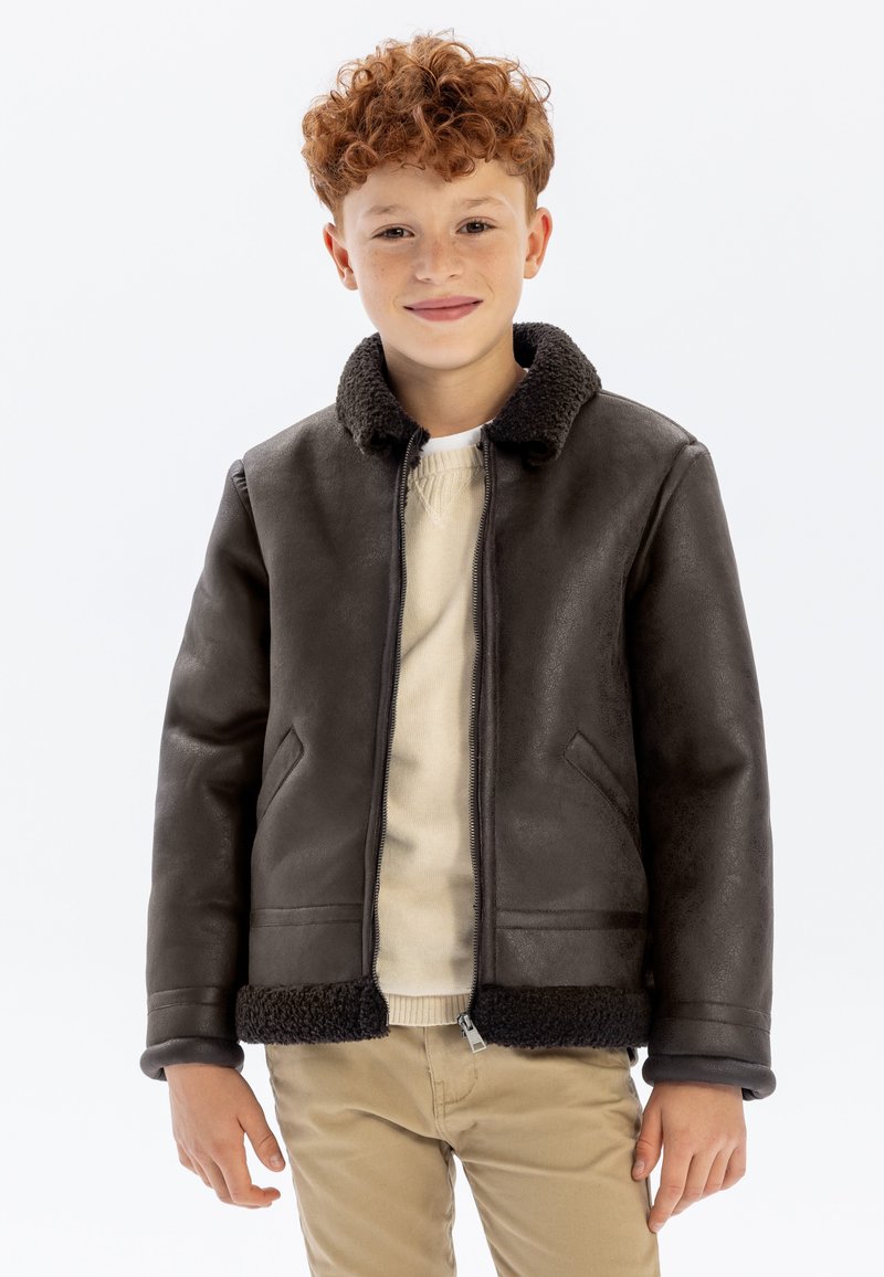 Veste bomber en faux cuir marron foncé avec un col en fourrure, fermeture zippée et deux poches avant. Portée sur un pull crème.