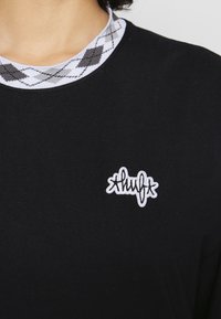 T-shirt preto com um colarinho argyle branco texturizado e um logotipo bordado branco que diz "Huf" na área do peito.