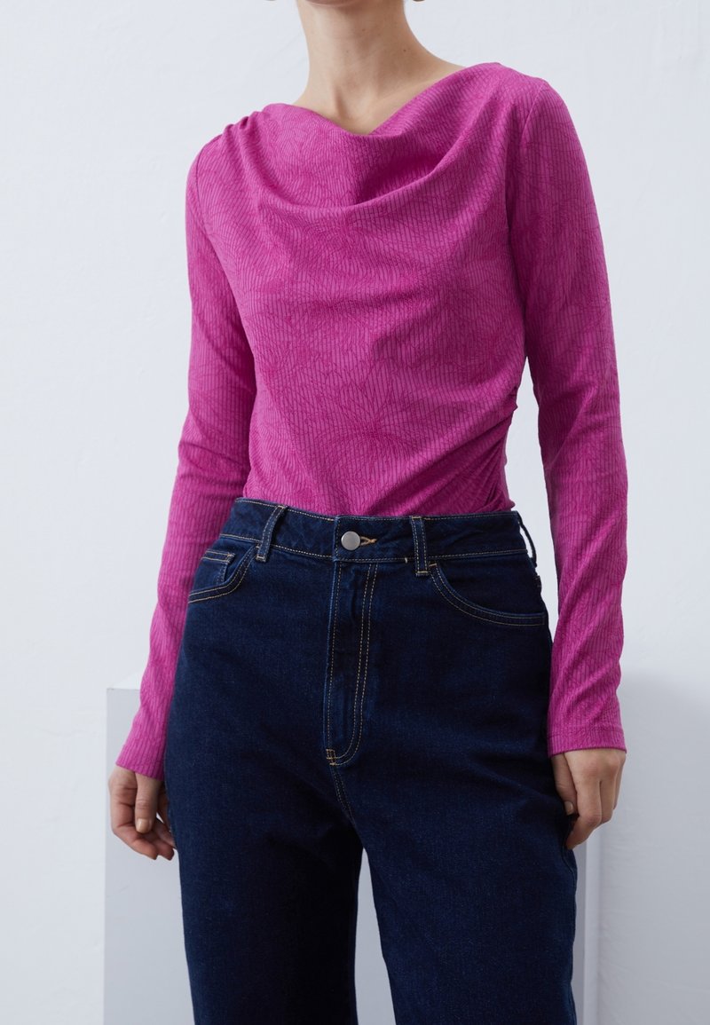 Enganliegendes langärmliges Oberteil aus leuchtend pinkfarbenem Rippstoff mit einem drapierten Ausschnitt. Kombiniert mit dunkelblauen, hochgeschnittenen Jeans.