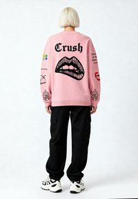 Felpa rosa con una grafica di labbra nere e la scritta "Crush" sul retro. I pantaloni cargo neri e le sneakers completano l'outfit.