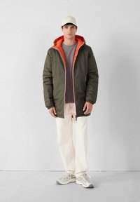 Giubbotto puffo verde oliva con interno arancione e chiusura a zip, indossato sopra una maglietta grigia e abbinato a pantaloni bianchi e sneakers chiare.