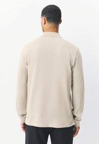 Vaaleanbeige pitkähihaispaita rentoutuneella istuvuudella, jossa on klassinen kaulus ja pehmeä, sileä kangas. Ei näkyviä kuvioita tai metalliosia.