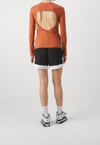 Långärmad topp i rostfärg med en stor ryggöppning, i kombination med svarta shorts med vit kant, och sportskor.