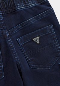 Jeans in denim scuro con un patch triangolare del logo "Guess" sulla tasca posteriore, con cuciture visibili e una superficie liscia.