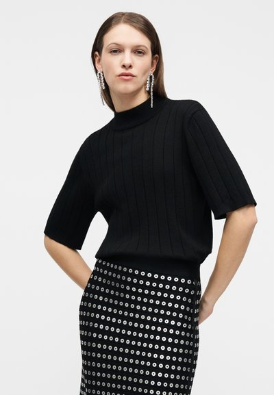 Γυναίκα που φοράει μαύρο ribbed μπλουζάκι με κοντά μανίκια, μαύρη φούστα με ασημένια μοτίβα από κρίκους και μακριά ασημένια κρεμαστά σκουλαρίκια, ποζάροντας με τα χέρια στη μέση.