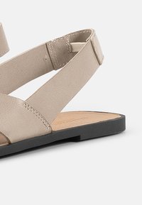 Beige skinnsandaler med kryssende stropper, flat svart såle og innersåle i naturlig tre-tone. Glatt tekstur og minimalistisk design.