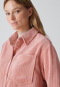 Pink corduroy shirt with a collar, featuring vertical ribbed texture, snap buttons, and a chest pocket. Smooth fabric in a relaxed fit.