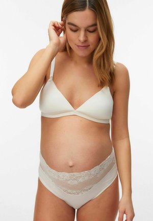 Schwangere Frau trägt weißen Schwangerschafts-BH und Spitzenunterwäsche, schaut nach unten mit der Hand nahe dem Ohr, vor einfarbigem Hintergrund.