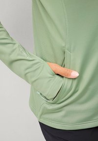 Světle zelený dlouhý rukáv top s měkkou texturou, úzkým střihem, boční kapsou a nařasením na zápěstí. Viditelné švy.