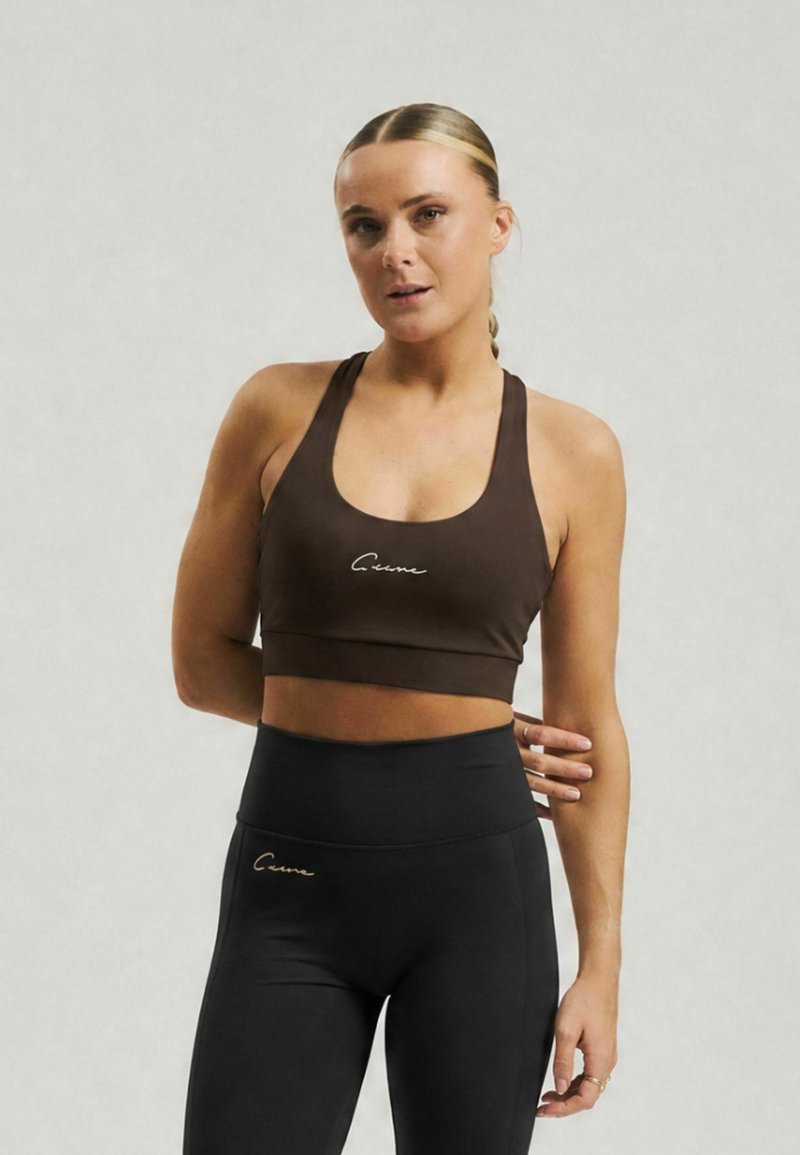 Femme aux cheveux blonds en tresse, portant un soutien-gorge de sport marron et un legging noir taille haute, debout devant un fond clair uni.