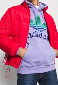 Röd puffjacka med hög krage och elastiska ärmar, över en lavendelfärgad hoodie med en blå och grön randig logotyp. Blå jeans.