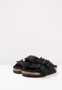 Chinelo preto de slip-on com textura peluda, palmilha de cortiça e duas tiras ajustáveis com fivelas pretas. Sola de borracha grossa.