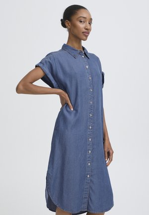 Femme debout, main sur la hanche, portant une robe en jean à manches courtes avec des boutons, sur un fond clair uni.