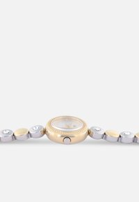 Montre en métal doré et argenté avec un cadran rond, des accents en cristal et une lunette texturée. Le bracelet est composé de maillons lisses et facettés alternés.