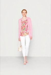 Mulher vestida com um blazer rosa, uma blusa floral, calças brancas cropped e sandálias com tiras, segurando uma mala creme, de pé contra um fundo claro.