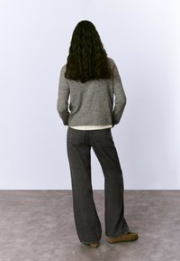 Mujer con cabello rizado largo, vistiendo un suéter gris, jeans anchos negros y zapatos mocasines marrones, de pie con las manos en los bolsillos.
