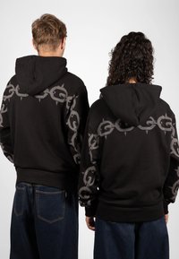 Schwarze Kapuzensweatshirts mit einem grauen Kettenmuster und Stacheln an den Schultern. Beide tragen lockere Jeans.