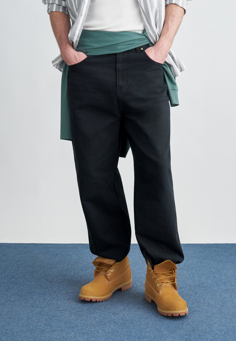 Μαύρο φαρδύ παντελόνι, συνδυασμένο με πράσινο πουκάμισο δεμένο στη μέση, συμπληρωμένο από καφέ μποτάκια με κορδόνια, ανθεκτική σόλα και κίτρινες λεπτομέρειες.