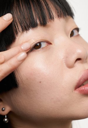 Primo piano di un volto con pelle liscia, trucco leggero e capelli neri lisci. Un dito tocca delicatamente la palpebra, accentuando la forma dell'occhio.