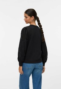 Svart sweatshirt med långa, puffiga ärmar som har intrikata hålmönster; kombinerad med ljusblå, löst sittande jeans.
