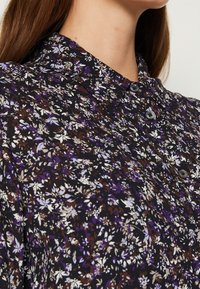 Femme portant un chemisier noir à motifs floraux violets, marron et blancs avec des boutons, montrée de la poitrine vers le haut avec col visible.