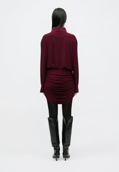 Raudonai burgundinio atspalvio suklijuota suknelė su apykakle, ilgomis rankovėmis ir suspausta tekstūra ties juosmeniu, derinama su juodais, kelius siekiančiais aukštakulniais batais.