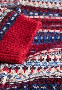 Gestrickter Pullover mit einem lebhaften roten Rippbündchen und mehrfarbigen Mustern in Rot, Blau und Weiß auf einem strukturierten Stoff.