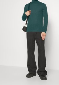 Tealfarbener Rollkragenpullover mit langen Ärmeln, kombiniert mit lockeren schwarzen Hosen und klobigen schwarzen Schuhen. Das Model hält eine kleine schwarz-weiße Tasche.
