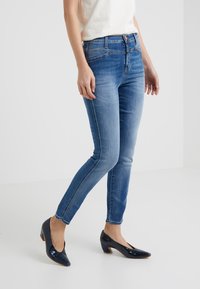 Pantalones vaqueros ajustados de mezclilla azul con un lavado desgastado, diseño de cinco bolsillos y costuras en contraste. Combinados con tacones brillantes en color marino.