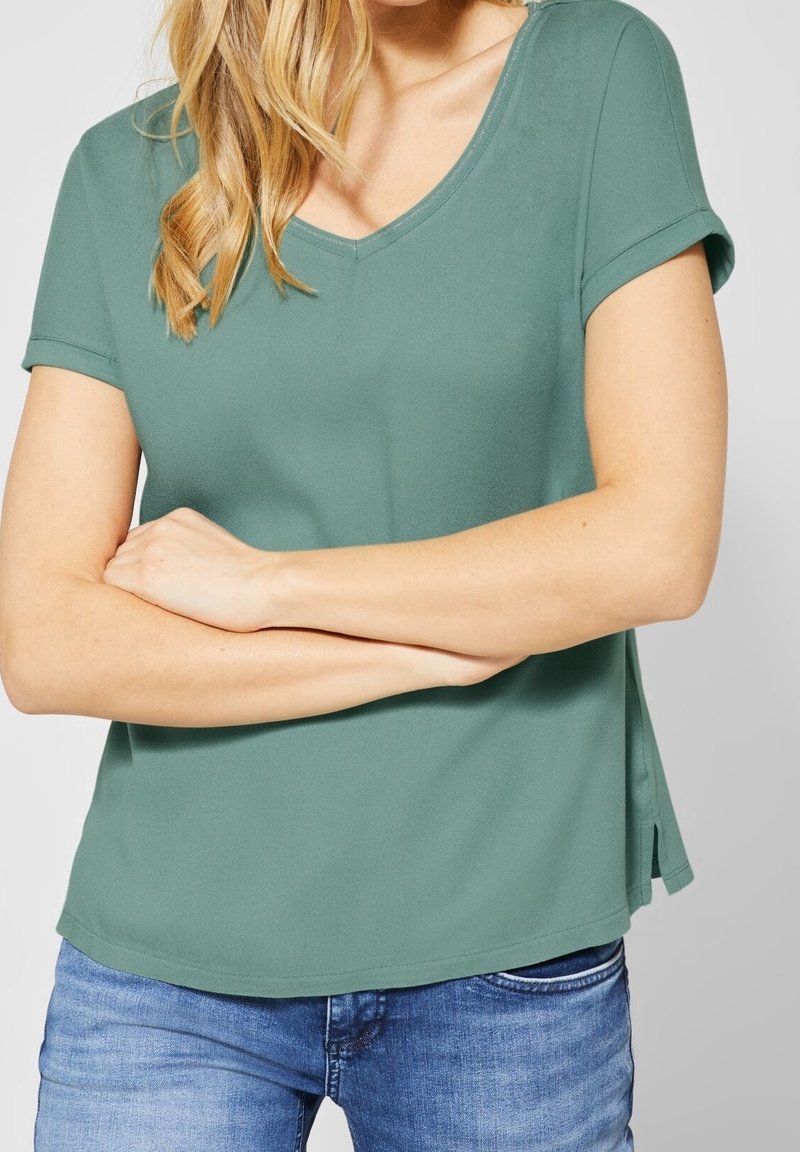 Grön kortärmad topp med V-ringning, tillverkad av ett mjukt tyg, med sidoklyftor, matchad med blå jeans.