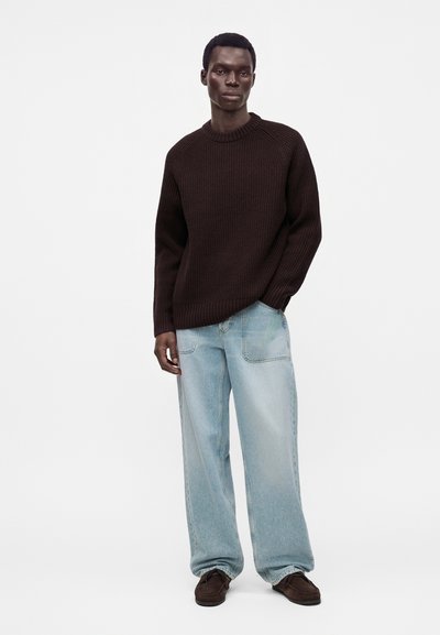Homme portant un pull en maille marron foncé, un jean bleu clair ample et des chaussures marron foncé, debout avec une main dans la poche.
