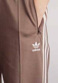 Rudos sportiniai kelnės su baltomis juostelėmis, šoniniu užtrauktuku kišene ir Adidas logotipu ant kairio šlaunies. Lygus audinys, pritaikytas kirpimas ir modernus dizainas.