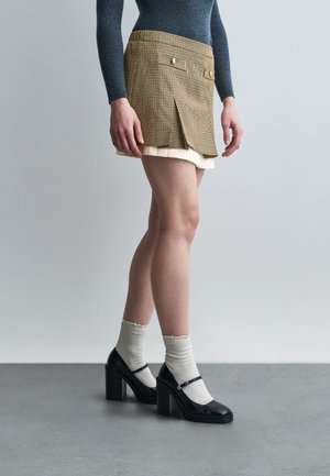 Osoba oblečená v vrstvenej béžovej károvanej sukni, tmavomodrom top s dlhými rukávmi, krémových ponožkách a čiernych blokových lodičkách Mary Jane stojaca na šedom podlahe.