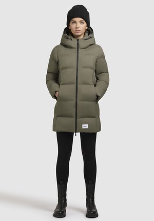 Khaki-puffer-takki, jossa on huppu, pitkät hihat ja vetoketjusuljenta. Varustettu kahdella sivutaskulla ja logopainatuksella helmassa. Yhdistetty mustiin legginsseihin ja saappaisiin.