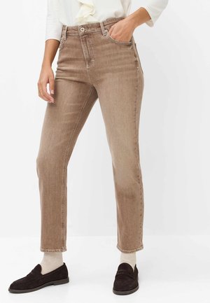 Femme portant un jean droit marron clair, des chaussettes blanches, des mocassins noirs et un top blanc à manches longues, une main dans la poche.