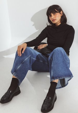 Jersey negro de cuello alto, jeans de pierna ancha azul con costuras prominentes, mocasines negros y bolso negro texturizado. Sentada sobre un fondo claro.