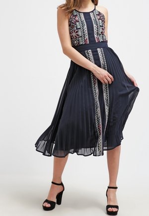 Vrouw draagt een mouwloze marineblauwe midi-jurk met plooien en geometrische borduurwerk, gecombineerd met zwarte enkellaarzen met hoge hakken en enkelbandjes.