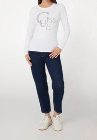 Biała koszulka z długim rękawem z srebrnymi cekinami z logo "GUESS", zestawiona z ciemnoniebieskimi dżinsami i białymi sneakersami. Luźny krój i prosty design.