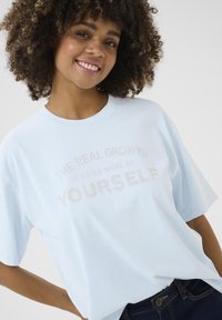 T-shirt surdimensionné bleu clair en tissu doux, avec un slogan texturé orné de rhinestones, centré à l'avant. Manches courtes.