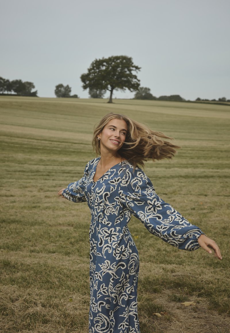 Junge Frau, die im Freien in einem blauen Blumenkleid mit langen Ärmeln dreht, auf einem grasbewachsenen Feld mit verstreuten Bäumen und bewölktem Himmel.