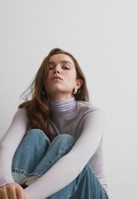 Haut à col roulé manches longues de couleur violet clair, associé à un jean en denim bleu. Le tissu semble lisse et ajusté, avec un léger éclat et des superpositions subtiles.