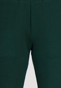 Donkergroene elastische broek gemaakt van een zachte, gestructureerde stof. Heeft een gladde tailleband en een aansluitend ontwerp. Geen zichtbare patronen of hardware.