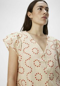 Vestido crema con patrón circular rojo, escote en V, mangas cortas con volumen y sutil detalle de botones en la parte delantera. Textura de tela ligera.