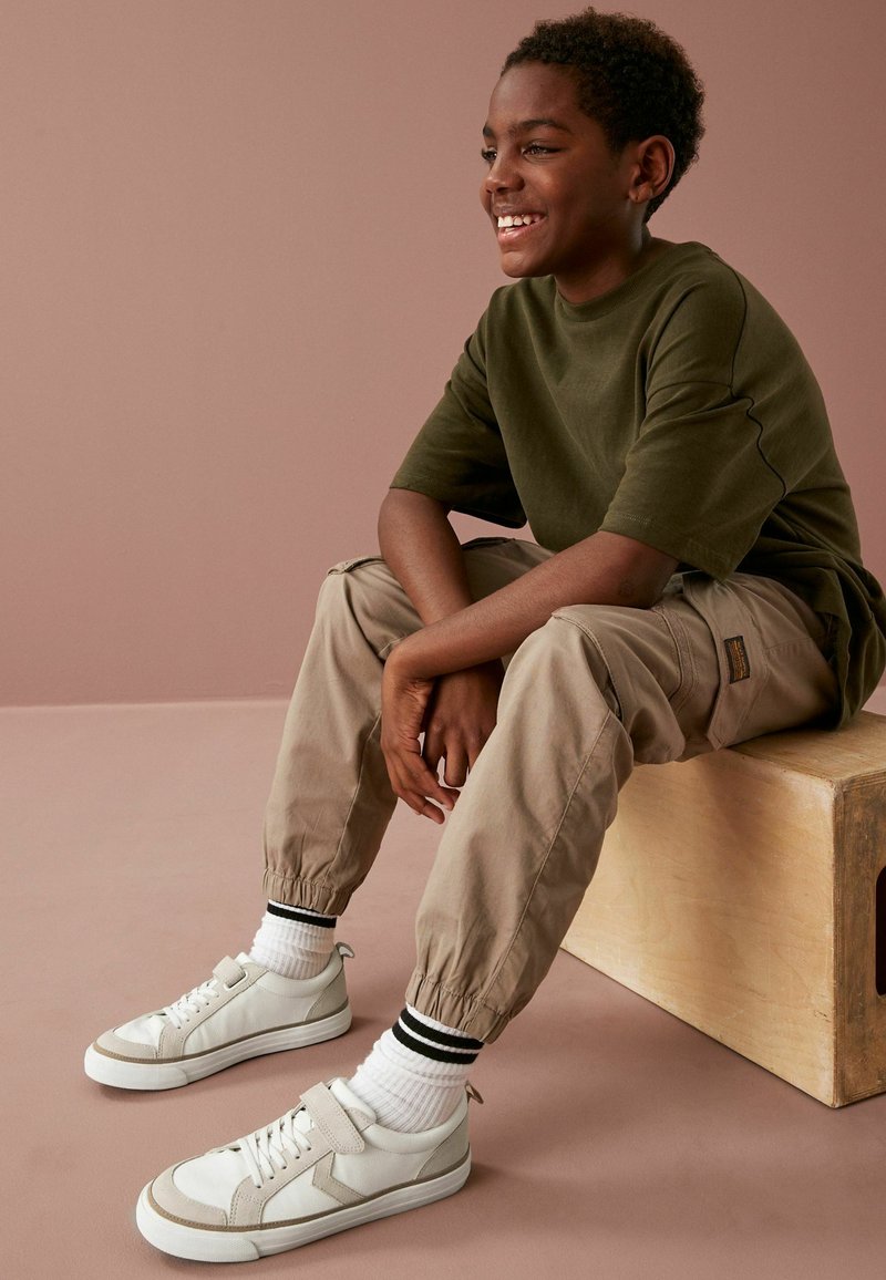 Khaki tapered byxor med elastiska manschetter, olivgrön t-shirt och vita sneakers med grå detaljer. Modellen sitter på en träblock.