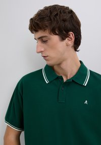 Polo de color verde oscuro con cuello de rayas blancas, tapeta de dos botones y un pequeño logo de calavera en el lado izquierdo del pecho. Mangas cortas, tejido texturizado.