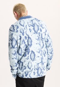 Ljusblå långärmad skjorta med ett blått ormtryckmönster. Har en krage och ribbade muddarna. Tyget verkar mjukt och texturerat.