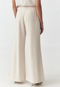 Pantalons larges en crème fabriqués à partir d'un tissu lisse, dotés d'une taille haute, de plis subtils et d'une coupe longue jusqu'au sol, associés à des talons.
