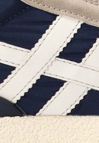 Zapatilla azul marino con detalles en cuero blanco y costura cruzada. Cuenta con una suela beige texturizada y un acabado suave en el material superior.