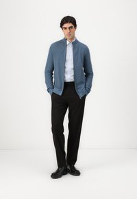 Blue zip-up knit cardigan over a light blue shirt, paired with black trousers and black boots. The cardigan has ribbed texture and a collar.